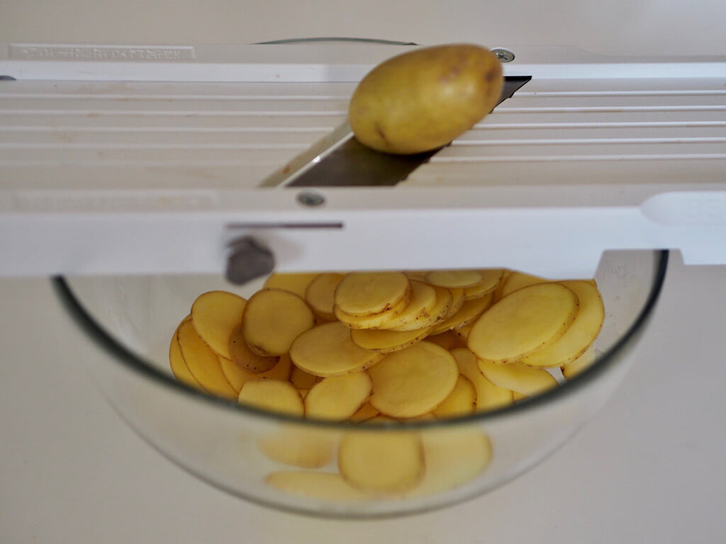 Siivutettuja perunaviipaleita lasikulhossa jonka päällä valkoinen mandoliini.